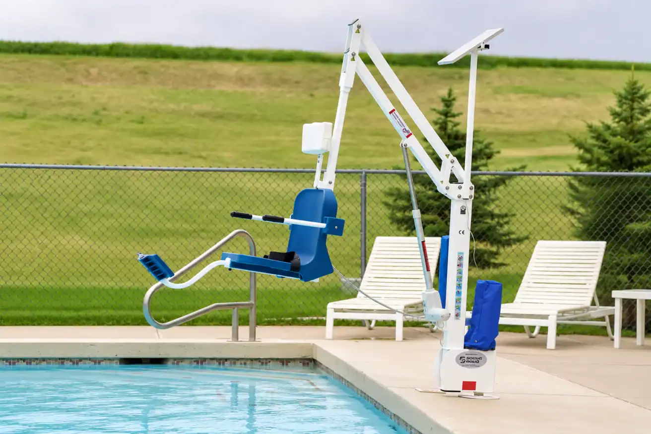A blue and white pool lift is positioned beside an outdoor swimming pool, with lounge chairs and a grassy field in the background.