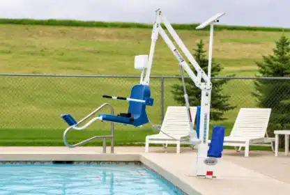 A blue and white pool lift is positioned beside an outdoor swimming pool, with lounge chairs and a grassy field in the background.