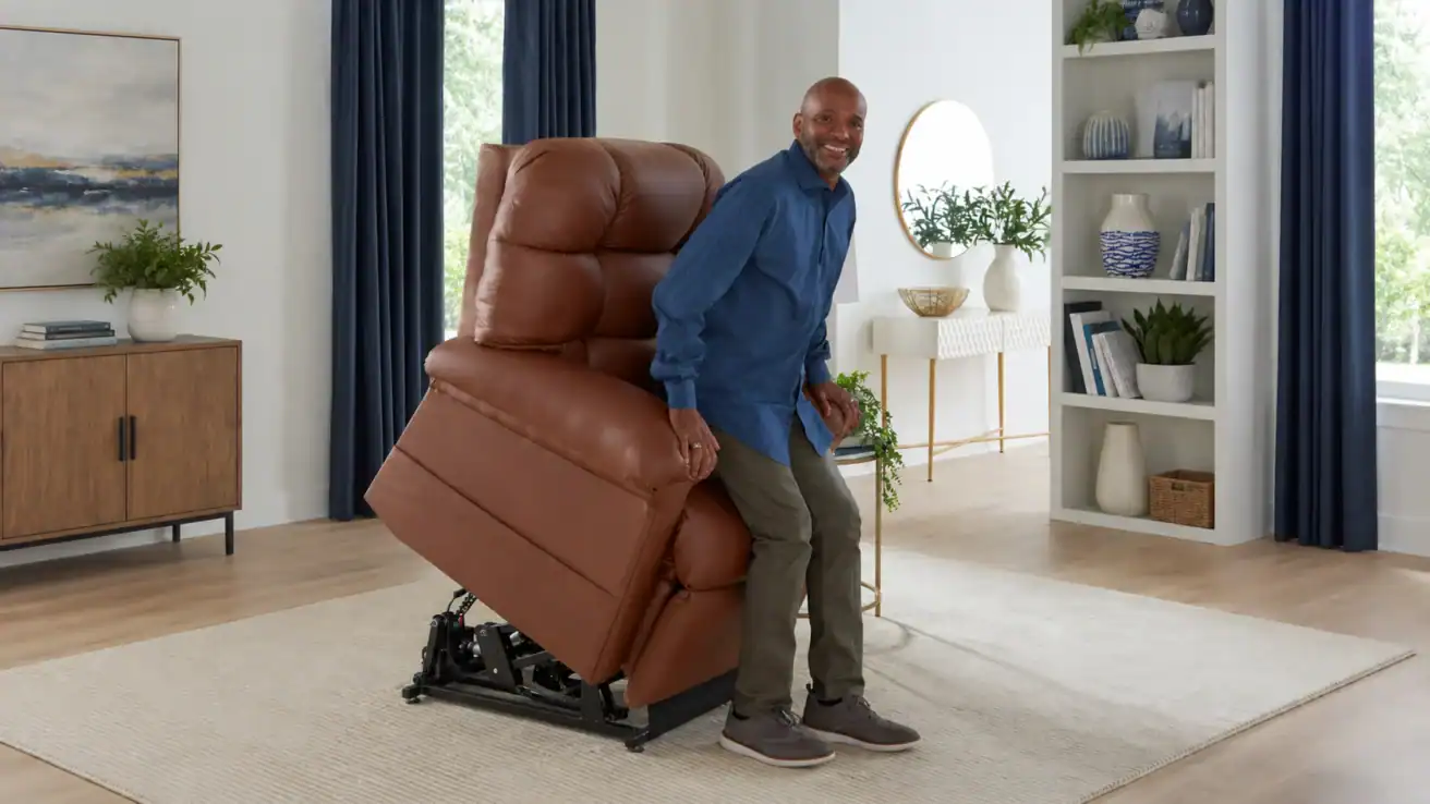 A man stands up from a brown leather lift chair in a living room with a beige rug, white walls, blue curtains, and shelves with books and decor.