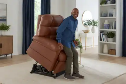 A man stands up from a brown leather lift chair in a living room with a beige rug, white walls, blue curtains, and shelves with books and decor.