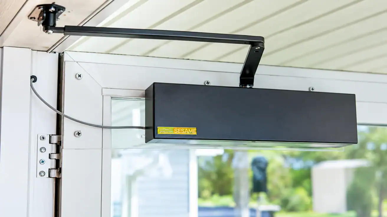 Automatic door opener device mounted on the top of a glass door, connected with a cable and support arm, with a yellow “SESAME” label visible.
