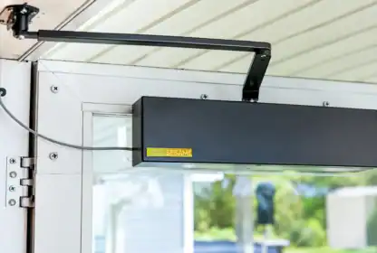 Automatic door opener device mounted on the top of a glass door, connected with a cable and support arm, with a yellow “SESAME” label visible.