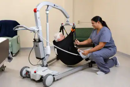 A healthcare worker kneels next to an elderly patient in a sling lift, assisting with patient transfer in a medical facility room.