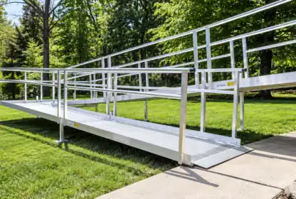 Aluminum wheelchair-accessible ramp with handrails leading from a concrete sidewalk to a grassy area, surrounded by trees and greenery.