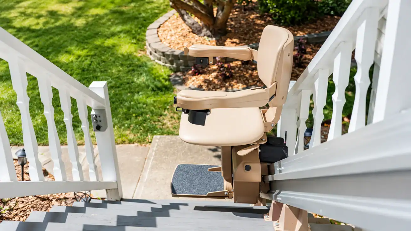 A beige stair lift is installed on an outdoor staircase with white railings, leading down to a garden with grass and landscaping.