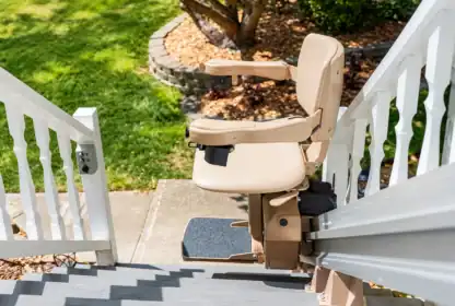 A beige stair lift is installed on an outdoor staircase with white railings, leading down to a garden with grass and landscaping.