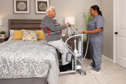 An elderly man uses a patient lift device next to a bed while a healthcare worker in scrubs assists him in a bedroom setting.