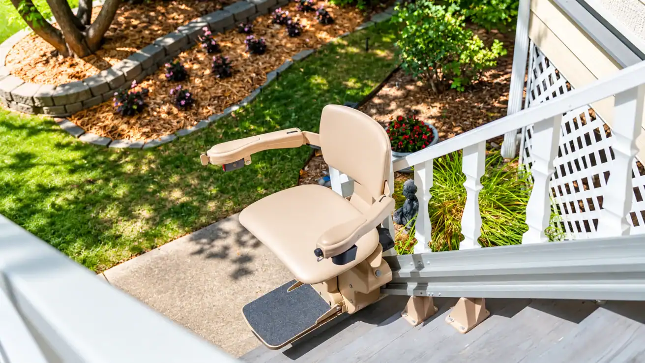 Outdoor stair lift chair installed on a staircase, overlooking a landscaped yard with flower beds and mulch, seen from the top of the stairs.