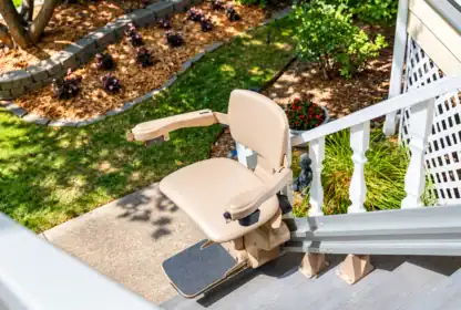 Outdoor stair lift chair installed on a staircase, overlooking a landscaped yard with flower beds and mulch, seen from the top of the stairs.