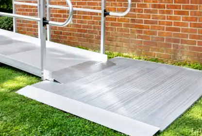 A metal wheelchair ramp with handrails leads from a grassy yard to a brick building.