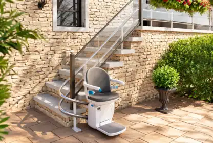 Outdoor stair lift installed alongside a short stone staircase by a brick house, surrounded by green shrubs and a potted plant on a patio.