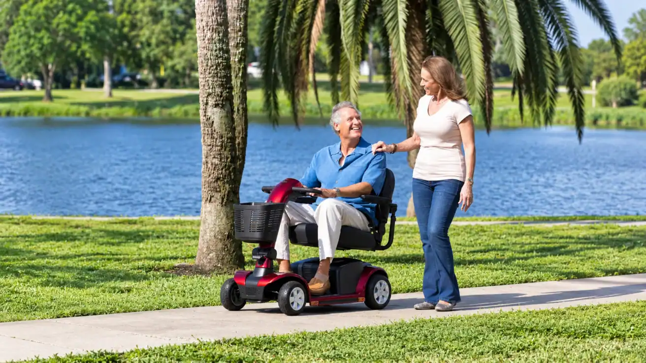 An older man uses a mobility scooter on a park path by a lake, smiling and talking with a woman walking beside him.