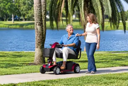An older man uses a mobility scooter on a park path by a lake, smiling and talking with a woman walking beside him.