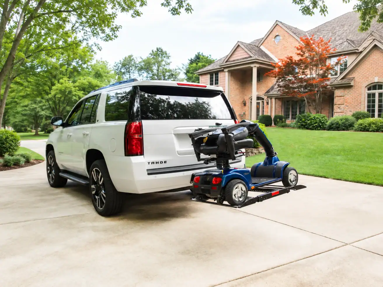 A white SUV with a mobility scooter secured to a rear-mounted lift is parked in the driveway of a brick house with trees and landscaping.
