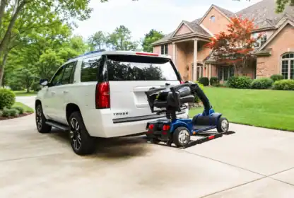 A white SUV with a mobility scooter secured to a rear-mounted lift is parked in the driveway of a brick house with trees and landscaping.