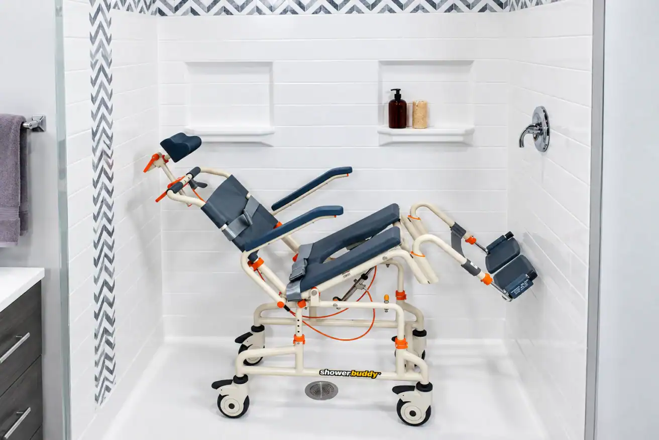 Adjustable shower chair with wheels and leg supports placed inside a tiled walk-in shower with built-in shelves holding bottles and towels.
