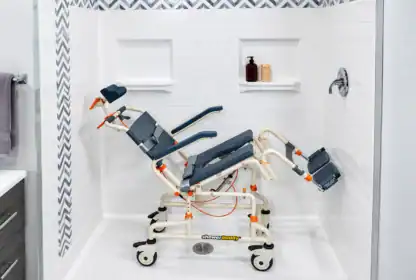 Adjustable shower chair with wheels and leg supports placed inside a tiled walk-in shower with built-in shelves holding bottles and towels.