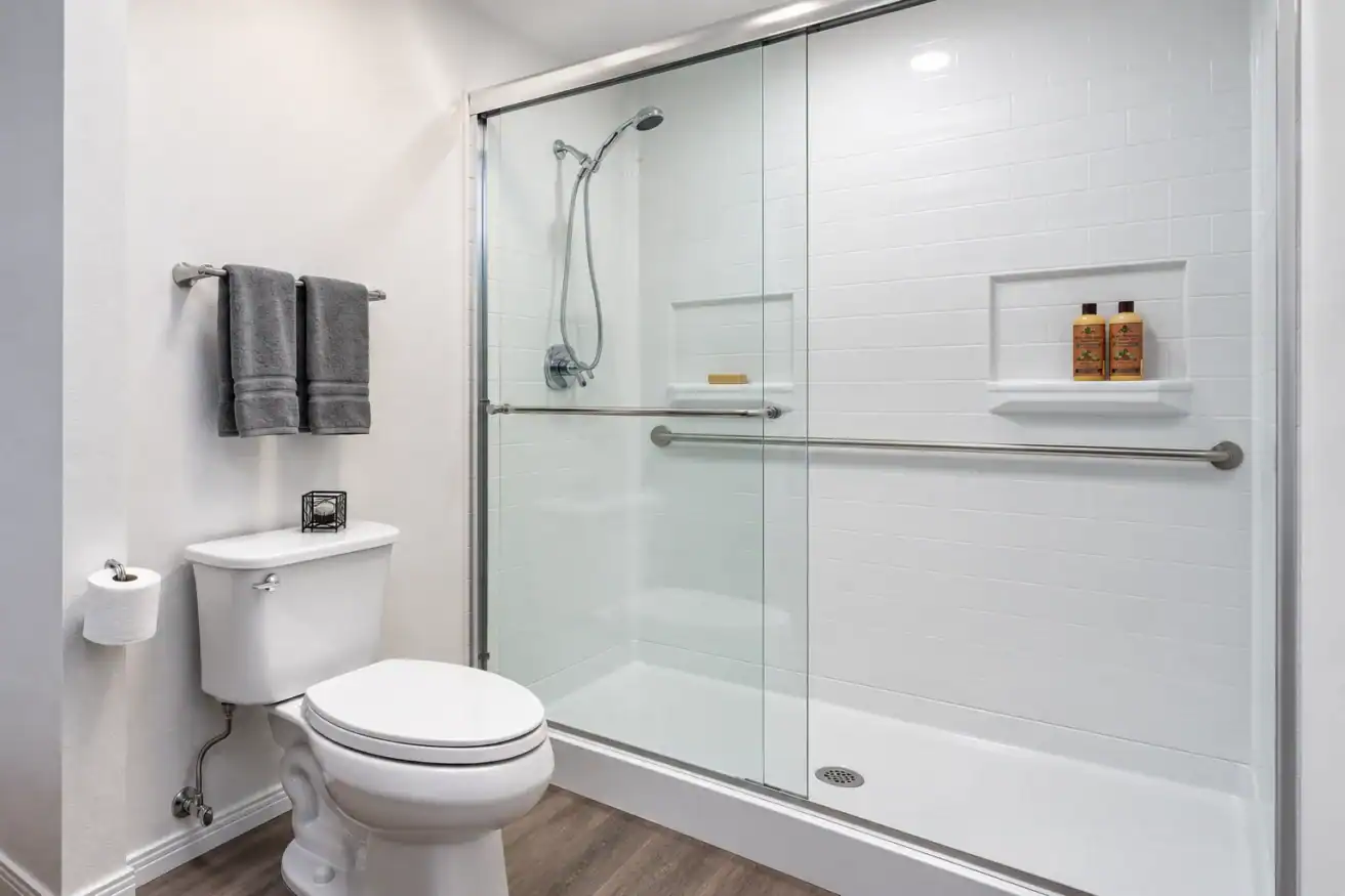 Modern bathroom with a glass-enclosed shower, white toilet, gray towels on a rack, toilet paper holder, and wood-look flooring. Two bottles and soap dish inside the shower.