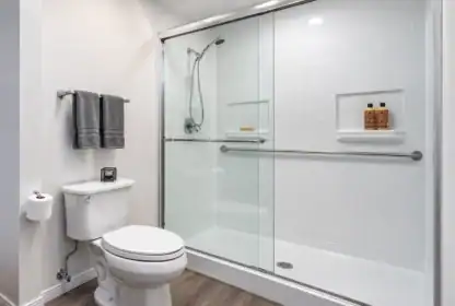 Modern bathroom with a glass-enclosed shower, white toilet, gray towels on a rack, toilet paper holder, and wood-look flooring. Two bottles and soap dish inside the shower.