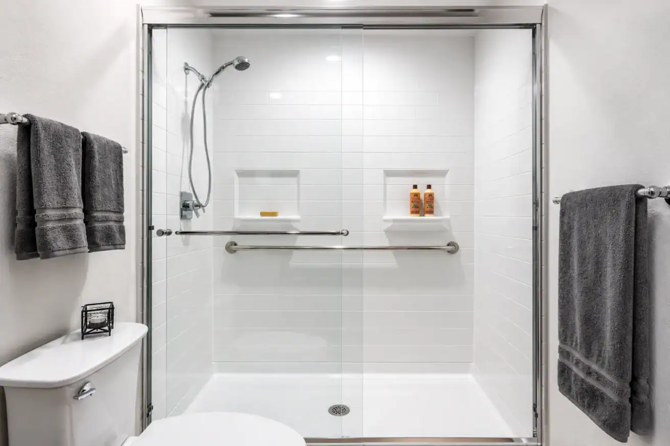Modern bathroom with a glass-enclosed shower, white tile walls, two built-in shelves, chrome fixtures, and three dark gray towels on racks.