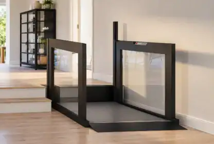 A black wheelchair lift is installed beside a short set of stairs in a modern, sunlit room with wooden floors and white walls.