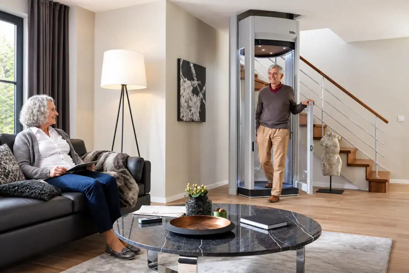 An older man exits a home elevator while an older woman sits on a sofa with a tablet in a modern living room.