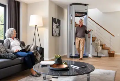 An older man exits a home elevator while an older woman sits on a sofa with a tablet in a modern living room.