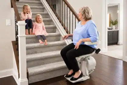 An older woman sits on a stair lift at the base of carpeted stairs, smiling at two young girls sitting on the steps in a home setting.