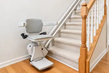 A motorized stair lift with a gray seat and armrests is installed along a carpeted staircase next to a wooden railing in a home.