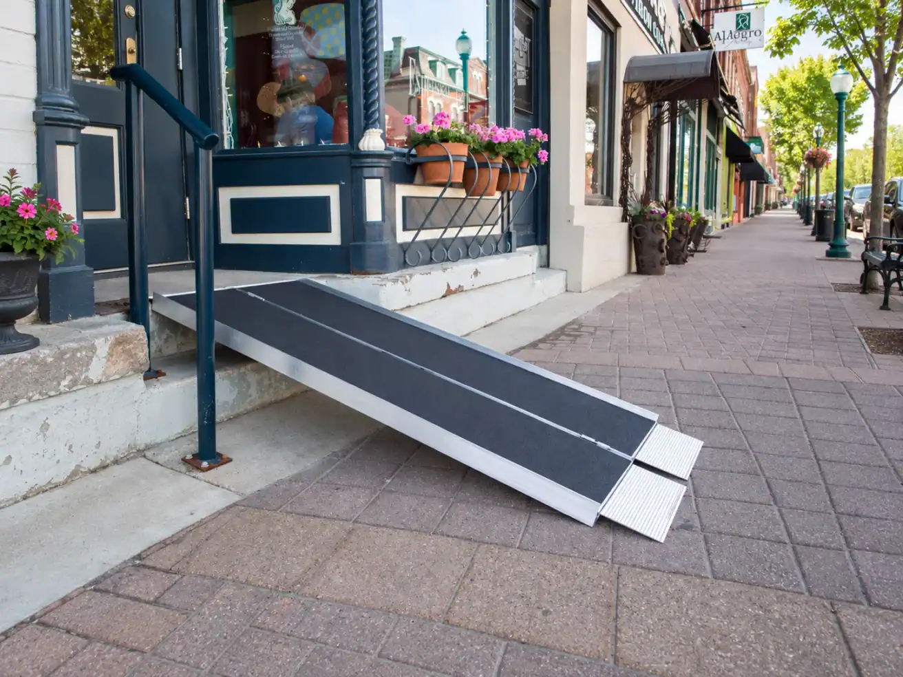 A portable wheelchair ramp leads from a storefront entrance down to the sidewalk on a quiet city street lined with shops and potted flowers.