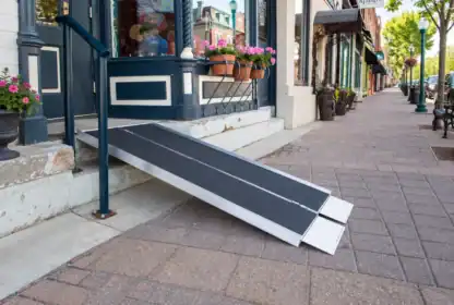 A portable wheelchair ramp leads from a storefront entrance down to the sidewalk on a quiet city street lined with shops and potted flowers.