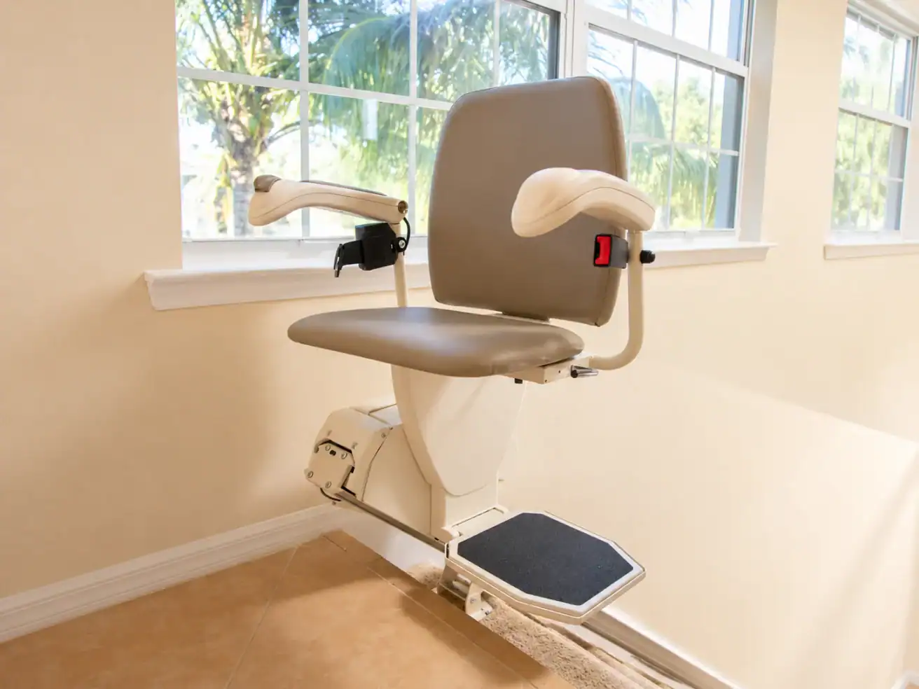 A beige stair lift chair with armrests and a footrest is installed next to a window in a bright room.