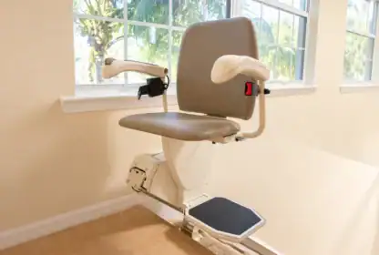 A beige stair lift chair with armrests and a footrest is installed next to a window in a bright room.