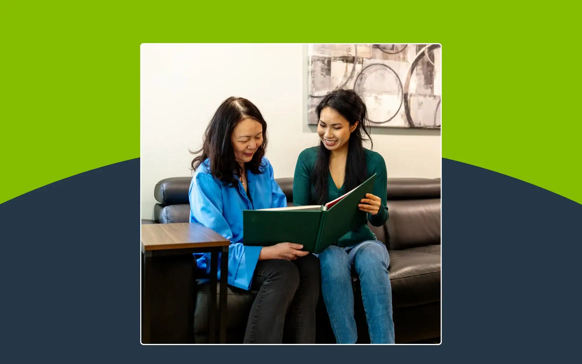 Two women sitting on a couch look at an open green book together, with a table on the left and abstract art on the wall behind them.