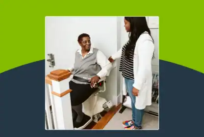 A caregiver standing next to an older woman seated on a stairlift.