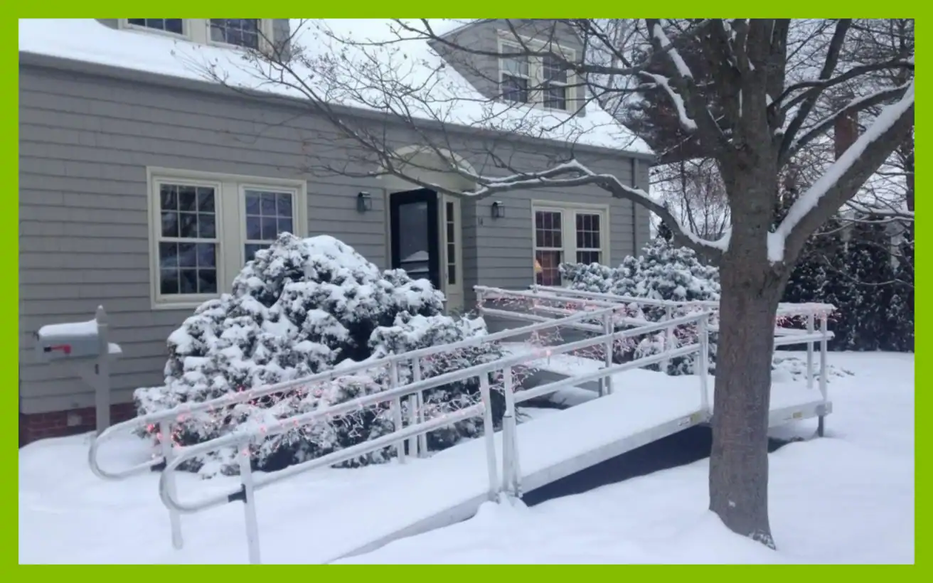A metal wheelchair ramp leading to the front door of a gray house covered in snow.
