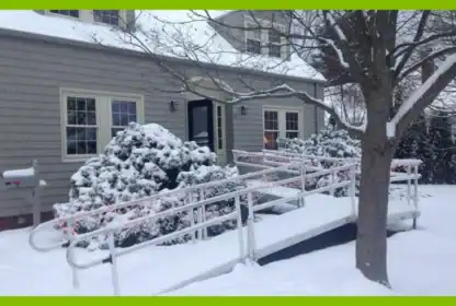 A metal wheelchair ramp leading to the front door of a gray house covered in snow.
