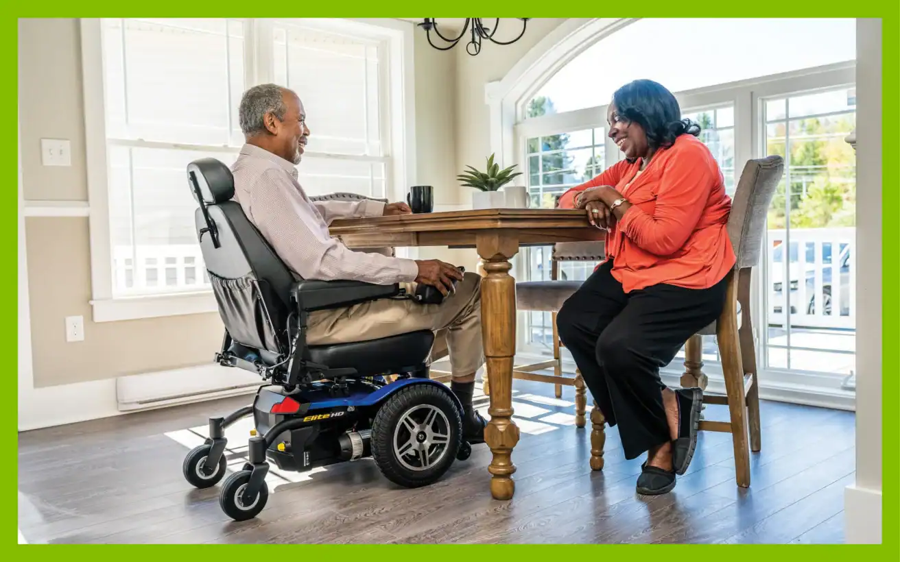 A man in a power wheelchair and a woman sit across from each other at a dining table, smiling and talking.