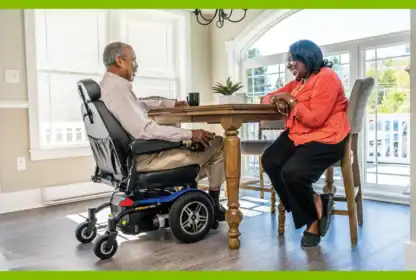 A man in a power wheelchair and a woman sit across from each other at a dining table, smiling and talking.