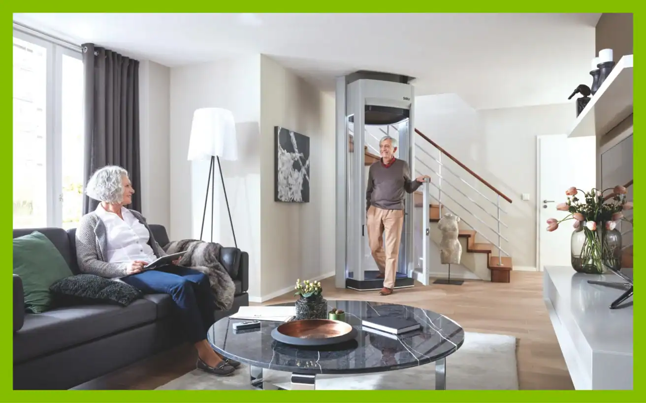 A man steps out of a residential elevator in a living room where a woman sits on a sofa.