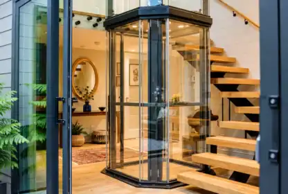 A modern glass elevator is installed next to a wooden staircase in a contemporary home interior with glass doors and a decorated entryway.