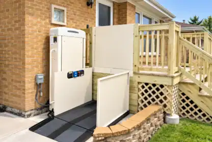 A vertical platform wheelchair lift installed next to a wooden deck and brick house.