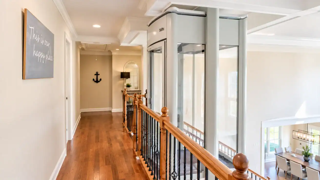 A Stiltz residential elevator installed in a hallway next to a wooden staircase.