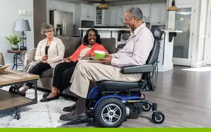 An older man in a blue power wheelchair holds a green bowl while sitting with two women in a living room.