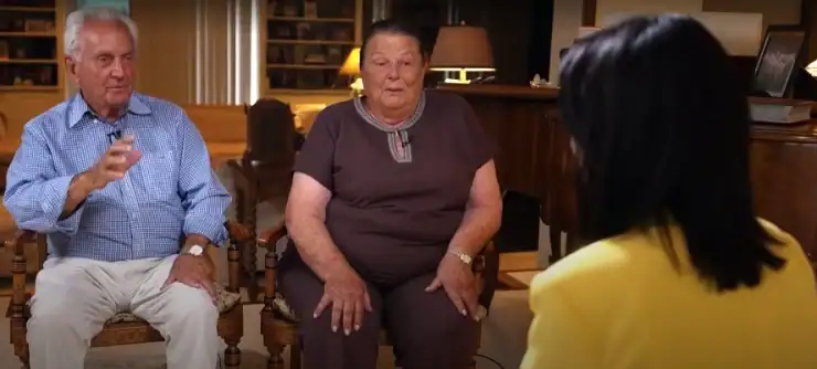 An older man and woman sitting in chairs during an interview, facing a person whose back is to the camera.